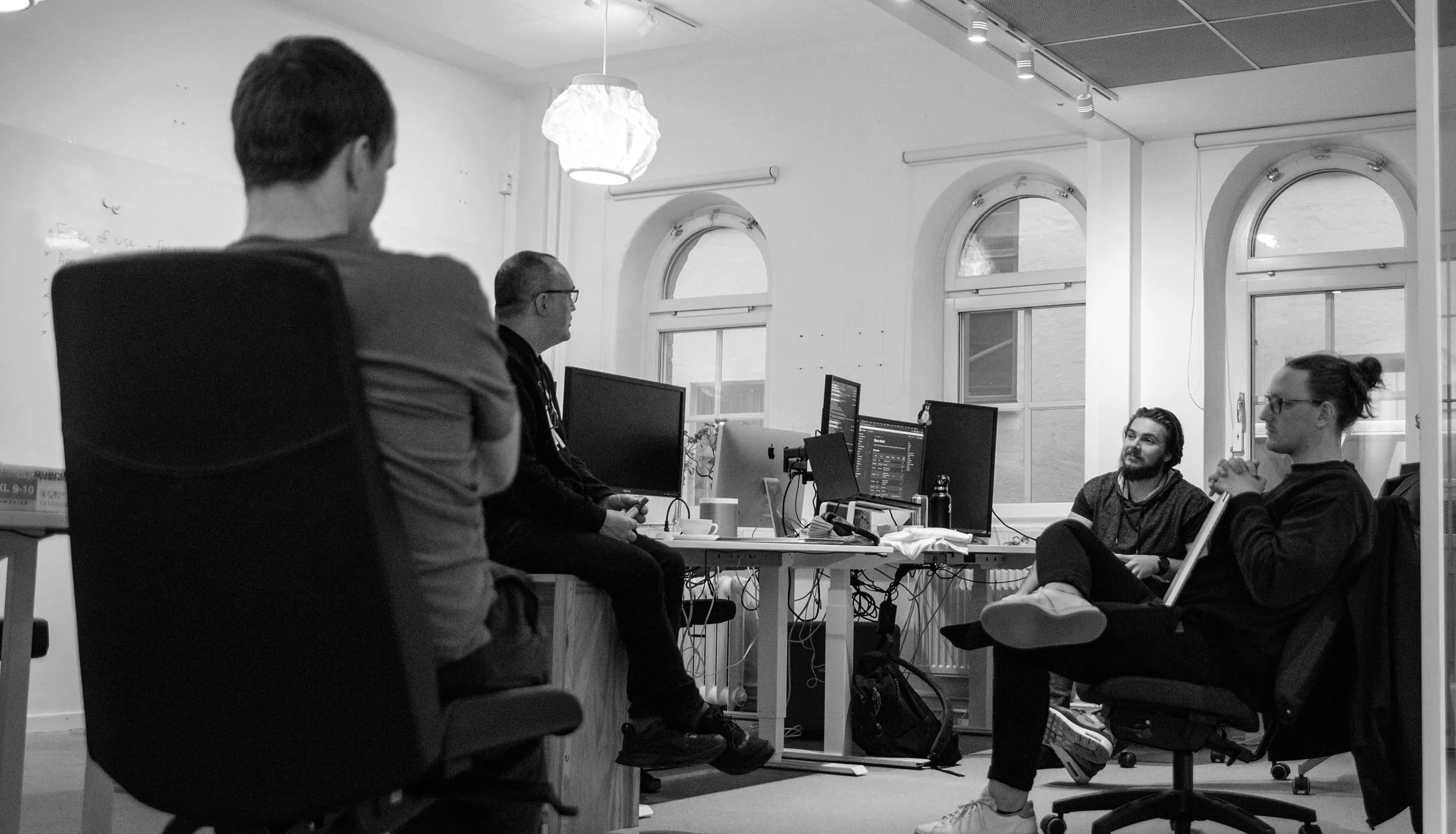 Four people engaged in discussion in an office, sitting around desks with computers and large arched windows in the background.